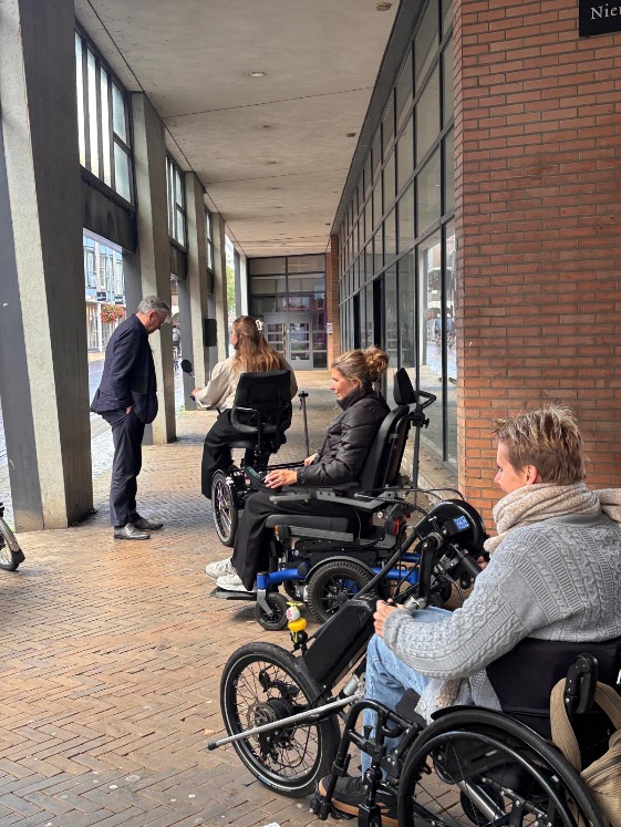 Drie vrouwen in rolstoelen en een man in pak staan onder een overdekte winkelgalerij met bakstenen gevel en klinkerbestrating.