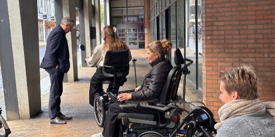 Drie vrouwen in rolstoelen en een man in pak staan onder een overdekte winkelgalerij met bakstenen gevel en klinkerbestrating.