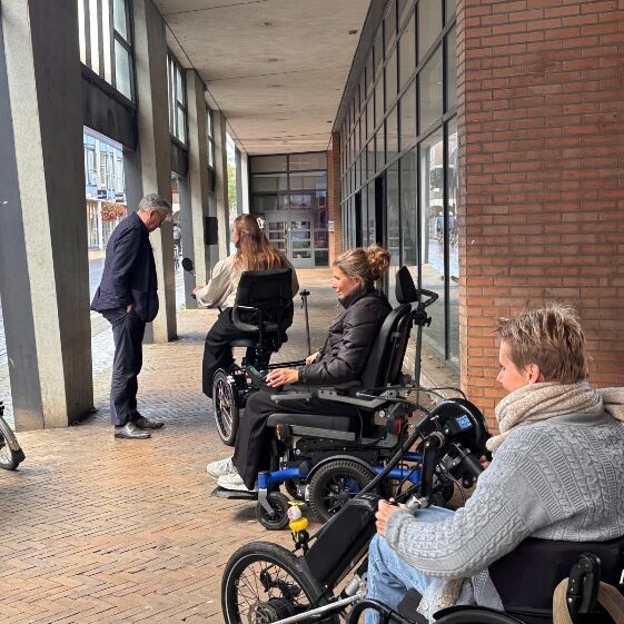 Drie vrouwen in rolstoelen en een man in pak staan onder een overdekte winkelgalerij met bakstenen gevel en klinkerbestrating.